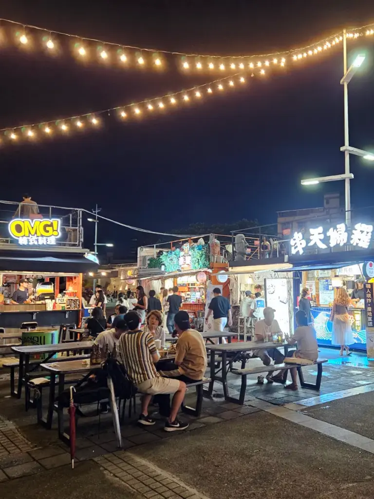 People sitting at picnic tables enjoying food and drinks at the Dadaocheng Wharf Container Market, illuminated by warm overhead string lights.
