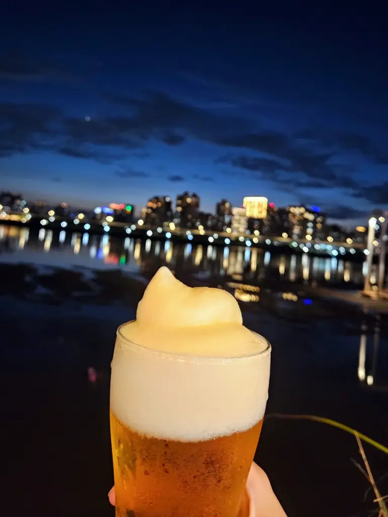 A close-up of a frosty glass of beer with a thick head of foam, held up against a blurred background of the Tamsui River and city skyline at night.