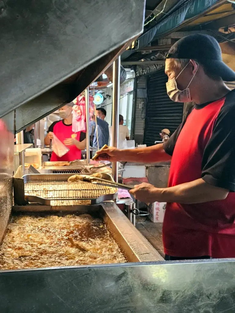 A vendor in a red shirt frying large chicken cutlets in a deep vat of hot oil at a night market stall.