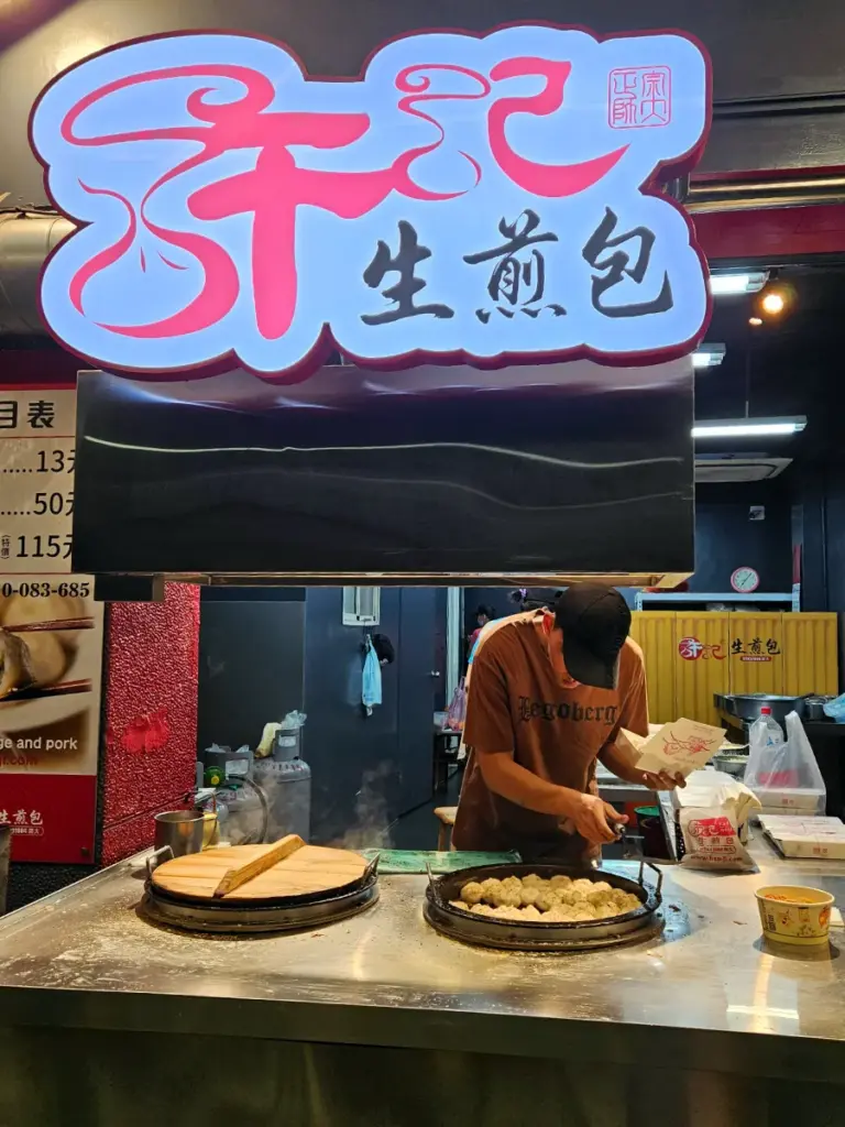 A vendor cooking a batch of pan-fried pork buns on a large circular flat-top grill under a neon sign.