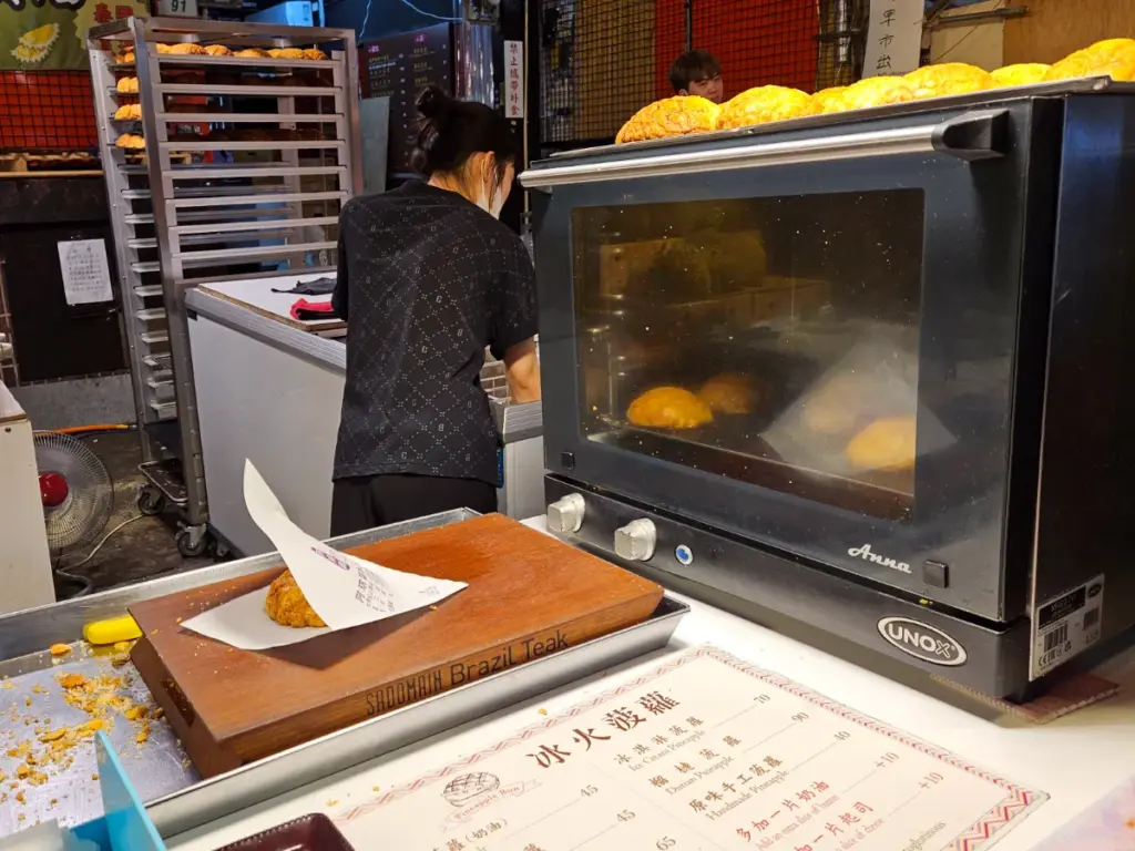 A worker tending to a commercial oven filled with freshly baked pineapple buns at a night market stall, with a menu displayed on the counter.