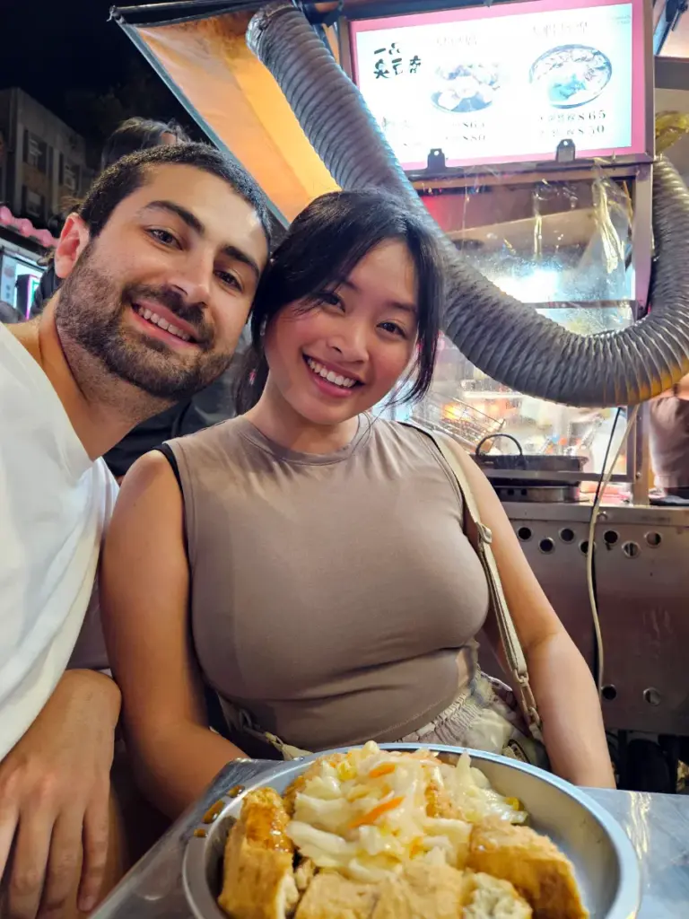 Tim and Zoe sitting at a night market stall with a plate of stinky tofu topped with pickled cabbage.