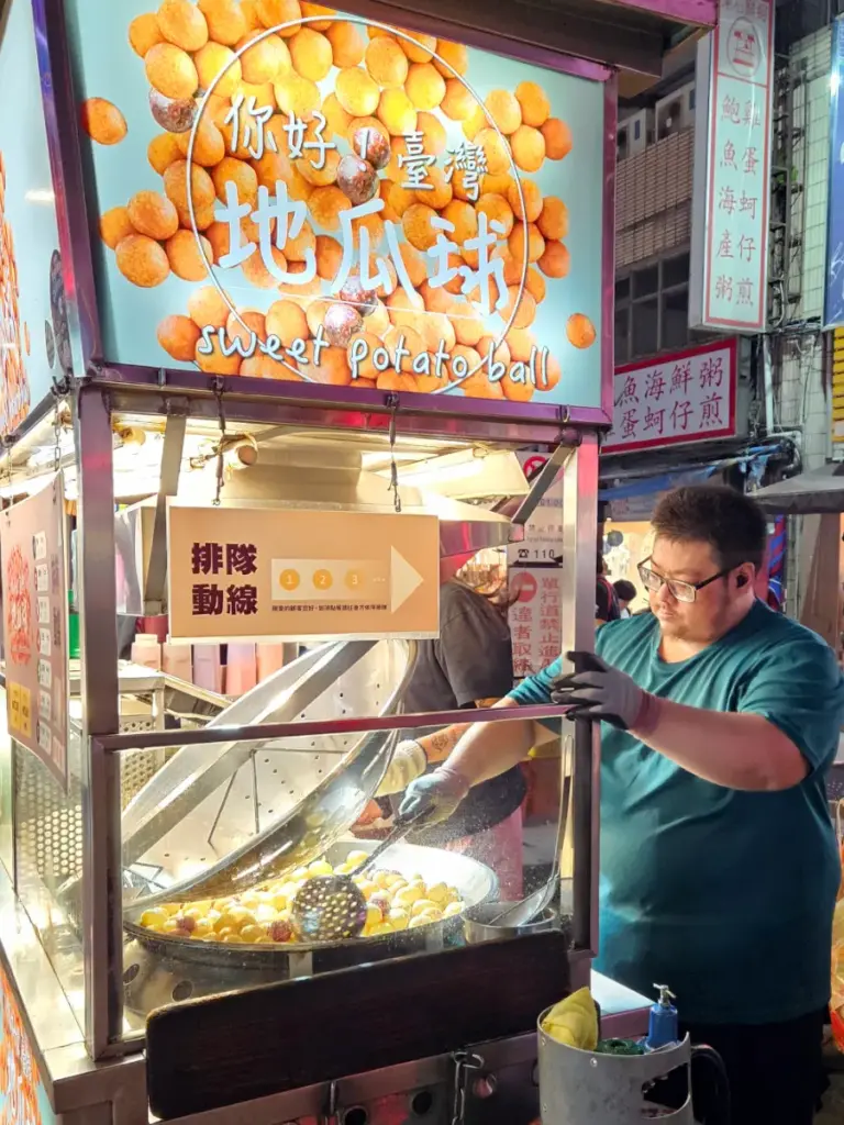 A night market vendor frying a large batch of sweet potato balls in a wok under a brightly lit stall sign.