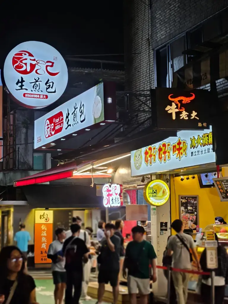 A bustling street view at Shida Night Market, one of the best Taipei night markets, showing the brightly lit signs for Hsu Ji Sheng Jian Bao and the adjacent Ho Ho Mei pineapple bun stall.