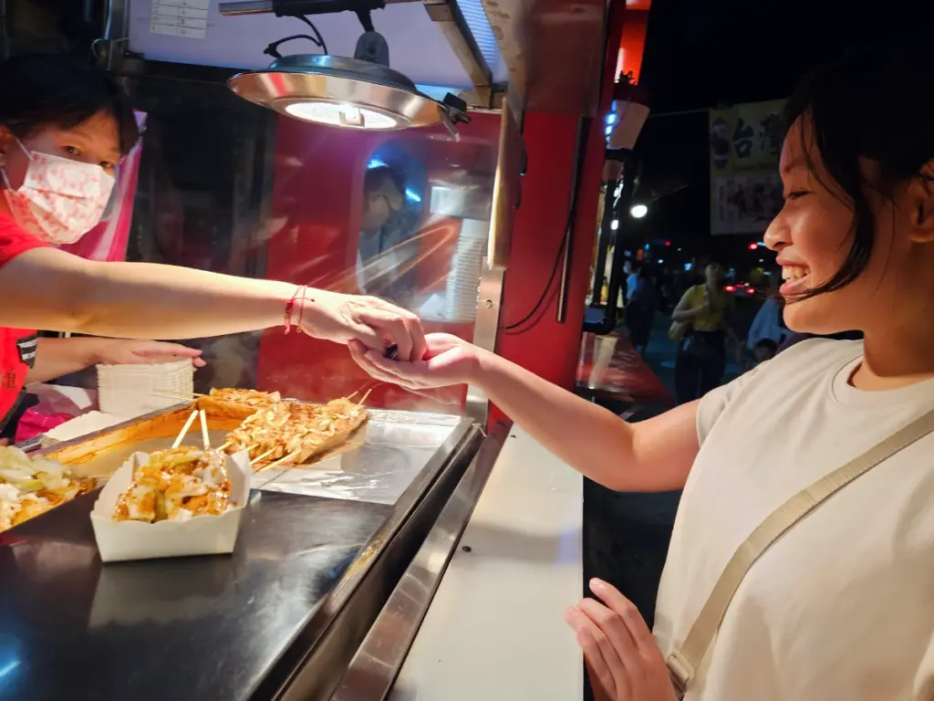 Zoe smiling as she buys a skewer of street food from a vendor at a bustling night market.
