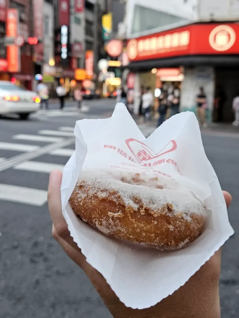 Tim holding a crispy milk donut in a paper wrapper against a blurred background of a city street.