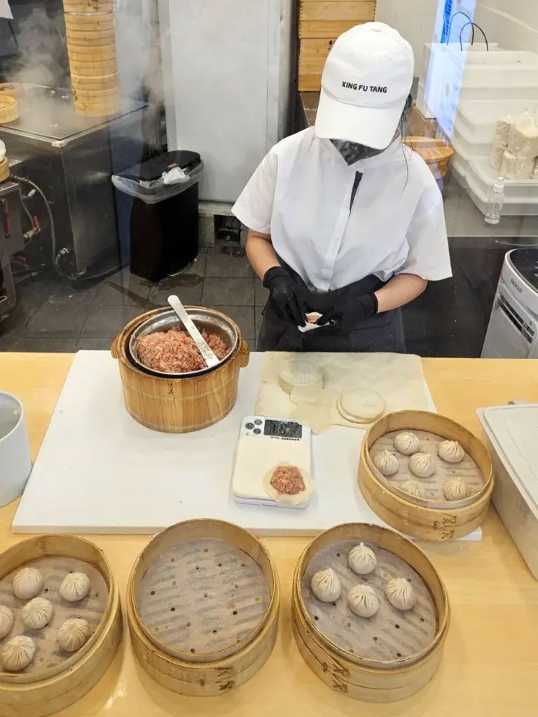 A worker wearing a white Xing Fu Tang cap preparing raw meat-filled xiaolongbao on a counter next to bamboo steamers.