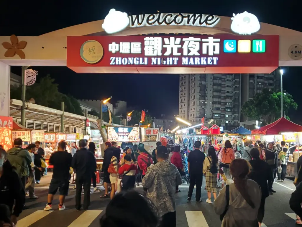 A crowd of people walking under the brightly lit welcome archway at the entrance of the Zhongli Night Market.